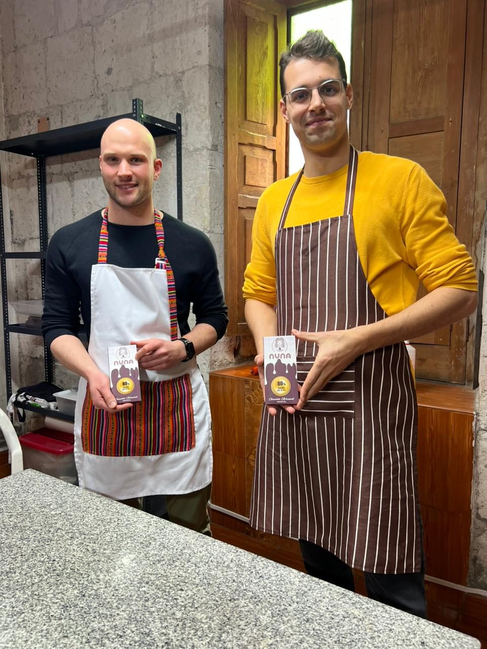 Two travelers in aprons smiling as they hold up the artisanal chocolate bars they just made in the workshop
